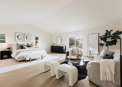 Spacious, modern bedroom with a king-sized bed, white bedding, and wooden accents. Features a sofa, armchair, and round coffee table. Decor includes framed art, a large mirror, and a potted plant. Neutral tones and ample natural light create a serene atmosphere.