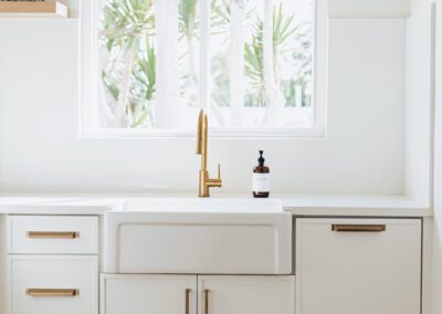 A clean, modern kitchen featuring white cabinets and a farmhouse sink with a brass faucet. Light pours in through the window, highlighting palm trees outside. The countertop has a soap dispenser, and a black wire basket sits on a shelf.