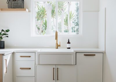 A modern kitchen with white cabinets and gold handles. A large farmhouse sink with a gold faucet is below a window, which shows a view of palm trees. A small potted plant and soap dispenser sit on the counter.