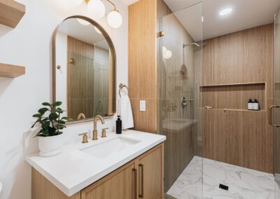 Modern bathroom with wooden accents, featuring a white sink with brass fixtures, a plant, and a soap dispenser. A rounded rectangular mirror is above the sink. The shower has a glass door, tiled walls, and built-in shelving. White and wood decor throughout.
