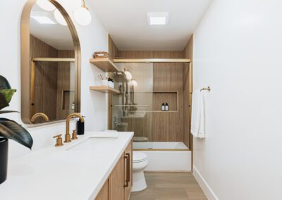 A modern bathroom with a white countertop and wooden cabinets. It features a golden faucet, a large mirror, a potted plant, shelves with toiletries, a towel rack, and a glass-enclosed bathtub with wood paneling. The lighting is warm and inviting.