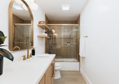 A modern bathroom with light wood cabinets and a white countertop. There is a round gold-framed mirror above the sink, and a glass-enclosed shower with wooden accents. A towel hangs on a gold towel bar, and a potted plant is on the counter.