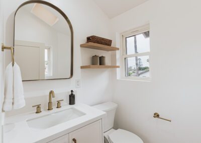 A minimalist bathroom with a white sink, brass faucet, and a large arched mirror. Two wooden shelves hold baskets and jars. A window provides natural light. Theres a white toilet and a brass toilet paper holder on the right wall.