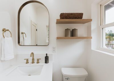 A modern bathroom with a white sink and brass fixtures. A large oval mirror hangs above, and a towel ring with a white towel is on the left. Wooden shelves on the right hold a basket and containers. A window lets in natural light.