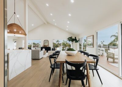 Modern open-concept living space with a high ceiling, featuring a dining table with black chairs. A living area with sofas is visible, and large windows overlook a patio and greenery. White walls and light wood floors enhance the airy feel.