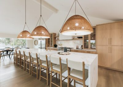Modern kitchen with a long marble island, surrounded by wooden bar stools. Three large woven pendant lights hang above. Light wood cabinetry and stainless steel appliances are in the background. Large windows bring in natural light.