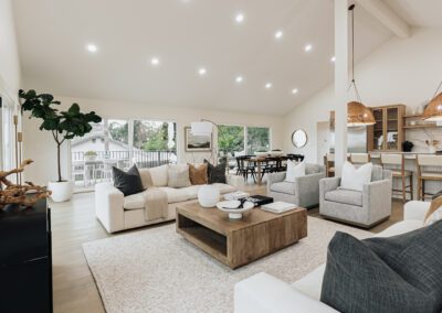 Modern living room with neutral tones featuring a large sectional sofa, plush armchairs, and a wooden coffee table on a light rug. Open kitchen with pendant lights and dining area visible in the background. Large windows provide natural light.