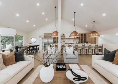 A modern, open-plan living and dining area with neutral tones. It features two sofas, a wooden coffee table with a decorative vase, wicker pendant lights above a kitchen island, and a dining table by a sliding glass door.