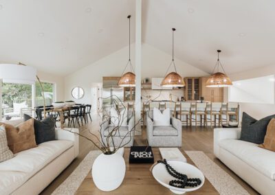 Spacious open-plan living room with a modern kitchen. Neutral tones dominate, with two large white sofas, a central wooden coffee table, and wicker pendant lights above a kitchen island. Large windows provide ample natural light.