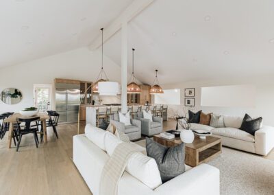 Spacious modern living room with vaulted ceilings, white sofas, and dark throw pillows. A wooden coffee table holds decorative items. Pendant lights hang above the kitchen area with stainless steel appliances and a dining table with black chairs.