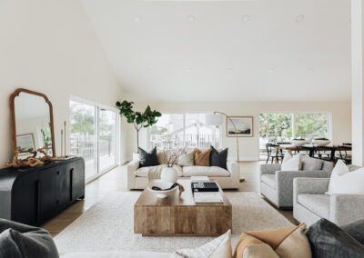 A spacious, modern living room with neutral tones, featuring white and gray sofas, a large wooden coffee table, and a black sideboard with a mirror. Large windows offer natural light and a view of greenery. A dining area is visible in the background.