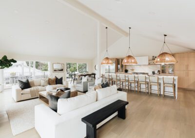 Spacious open-concept living area featuring a modern kitchen with white cabinets and wooden accents. Theres an island with bar stools, a large dining table, and a cozy seating area with sofas and a coffee table, all under high, vaulted ceilings.