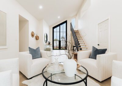 Modern living room with two beige armchairs and a round glass coffee table. The table holds decorative vases. A staircase with black railing leads upstairs, and large windows allow natural light to fill the space. Neutral tones dominate the decor.