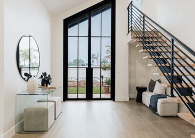 A modern, minimalist living space with light wooden floors, a black-framed floor-to-ceiling window, and a staircase with a black railing. A round mirror and decor sit on a glass table, and a cushioned seat is below the stairs.