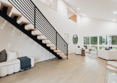 A modern living room with light wood flooring, a floating staircase with black railing, and a cozy seating area. Two chairs and a small table face a large window. A round mirror adorns the wall, and a cushioned bench sits below the stairs.