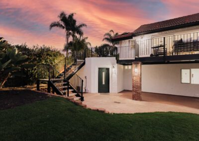 A two-story house with a sunset sky background. The house has an upper balcony with black railings, palm trees nearby, and a green front door. There are brick pillars on the lower level and a grassy yard in the foreground.
